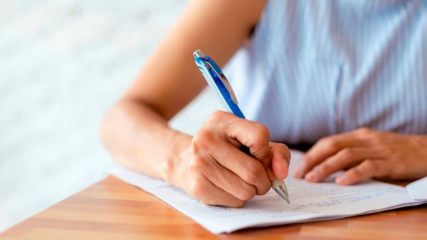 Eine Frau hält einen blauen Kugelschreiber in einer Hand und notiert etwas auf einem weißen Blatt. | Bild: mauritius images / Kittipong Satrinekarn / Alamy / Alamy Stock Photos