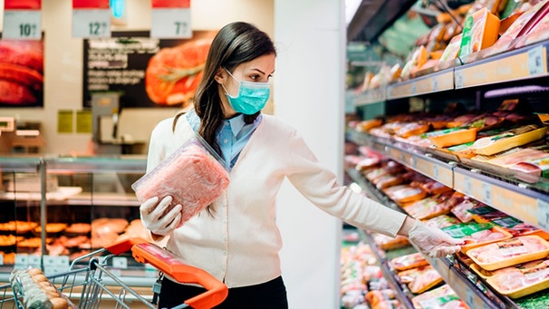 Junge Frau vor eingeschweisstem Fleisch im Supermarkt, sie trägt eine Maske und überlegt, welches Produkt sie kaufen soll. | Bild: mauritius-images
