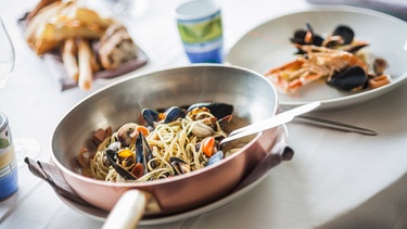 Eine Kupferpfanne steht auf dem Tisch. In der Pfanne und auf einem Teller liegen Spaghetti mit Muscheln.  | Bild: mauritius images / foodcollection 
